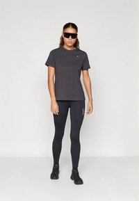 Nainen, joka on pukeutunut mustiin aurinkolaseihin, tummanharmaaseen t-paitaan, mustiin legginseihin, joissa on logo, ja mustiin lenkkareihin, seisoo valkoista taustaa vasten.