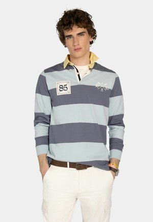 Joven con cabello rizado que lleva una camiseta de rugby de manga larga a rayas azul y gris, con cuello amarillo, pantalones blancos y cinturón marrón.