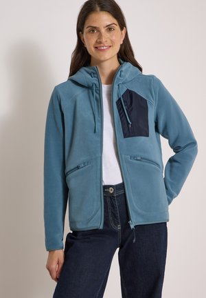 Femme portant une veste polaire bleue zippée avec une poche poitrine noire, chemise blanche et jeans foncés, souriante avec une main sur la hanche.