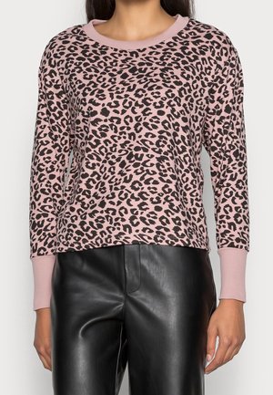 Frau trägt ein pinkfarbenes, langärmliges Leopardenmuster-Shirt kombiniert mit schwarzen Lederhosen, steht vor einem schlichten Hintergrund.