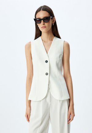Witte mouwloze top met V-hals, twee knopen aan de voorkant en een getailleerde pasvorm. Gepaard met bijpassende witte broek en zwarte cat-eye zonnebril.