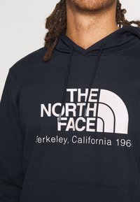 Muž s tmavými kudrnatými vlasy oblečený v tmavě modré mikině s kapucí, na které je v bílé barvě vytištěno logo "The North Face Berkeley, California 196".