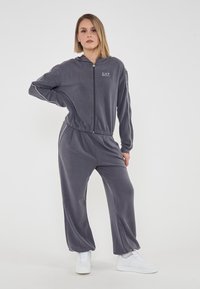 Tuta grigia con zip e cappuccio, dotata di vita e polsini elasticizzati. Tessuto morbido e liscio con strisce laterali a contrasto. Indossato con sneakers bianche.