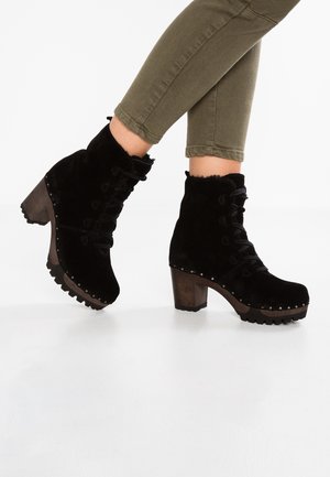 Sorte ankelstøvler i ruskind med snørebånd, træblokhæl og nitter, båret med opkrogede olivengrønne bukser.