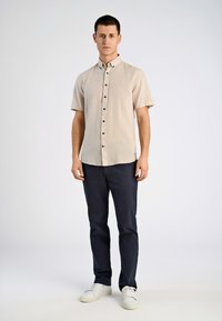 Camisa de botones de manga corta color beige con un acabado texturizado, combinada con pantalones azul marino oscuros y zapatillas blancas, de pie contra un fondo liso.