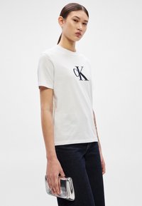 Valge puuvillane T-särk väikese tikitud "ck" logoga rinnal, kombineeritud tumedate teksadega ja hoides hõbedast metallist clutch-kotti.