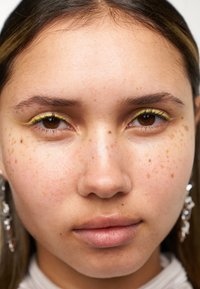 Nærbilde av et ansikt med lys brun hud og fregner. Grønn eyeliner fremhever øynene, supplert med klare dråpeøreringer og jevne lepper.