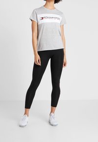 Šedé tričko s krátkým rukávem s bílým pruhem nesoucím nápis „TOMMY SPORT“, doplněné černými legínami a bílými sportovními botami.