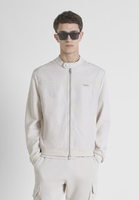 Chaqueta bomber de color beige claro con cremallera frontal, cierre de botones a presión y acentos texturizados. Combinada con pantalones a juego y gafas de sol oscuras.