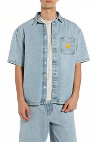 Camisa de ganga azul clara de manga curta com fecho de botão à frente, bolso no peito e etiqueta da marca amarela. Textura suave, silhueta descontraída.