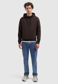 Jonge man met krullend haar, gedragen een donkerbruine hoodie met de tekst "PURE PATH", blauwe spijkerbroek en witte sneakers, staand tegen een lichte achtergrond.