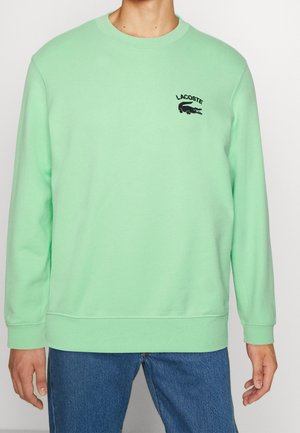 Homme portant un sweat-shirt à manches longues vert clair avec un logo Lacoste noir sur le côté gauche de la poitrine et un jean bleu, debout devant un fond uni.