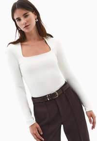 Donna che indossa una maglietta bianca a collo quadrato con maniche lunghe, pantaloni marrone scuro a vita alta e una cintura abbinata con fibbia dorata.