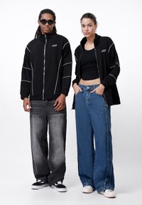 Black zip-up jacket with white accents, paired with gray baggy jeans. Model on left wears sunglasses; model on right has a black crop top.