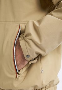 Jachetă bej cu fermoar, având un buzunar lateral cu fermoar alb, mansete elastice și un material texturat cu un model subtil în carouri.