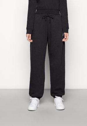Personne portant un pantalon de survêtement noir avec cordon et des baskets blanches, debout devant un fond clair uni.