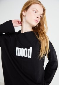 Crna majica dugih rukava s tiskanim riječima "mood" u velikim bijelim slovima na prednjem dijelu. Model nosi velike naušnice i ima dugu kosu.