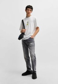 Hvid t-shirt med korte ærmer og gul og sort grafisk design, parret med grå denim jeans og sorte sneakers, og med en crossbody taske.