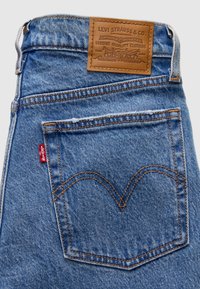 Τσέπη πίσω από μπλε τζιν Levi's με πορτοκαλί ραφές και καφέ δερμάτινο ταμπελάκι της μάρκας Levi Strauss & Co. πάνω από την τσέπη.