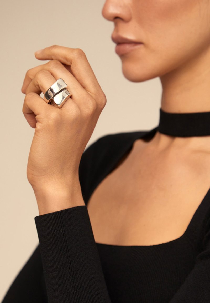 Anillo de plata con un diseño suave y redondeado que presenta dos bandas entrelazadas. El modelo lleva una blusa negra ajustada con un escote amplio.