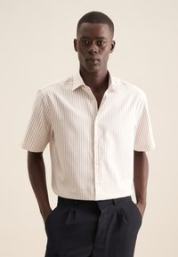 Camicia a maniche corte color beige chiaro con strisce bianche verticali, con bottoni sul davanti, colletto classico e vestibilità rilassata. Indossata con pantaloni blu navy.