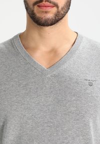 Šedý svetr s výstřihem do V vyrobený z pleteniny. Disponuje jemnou texturou a malým logem na levé části hrudi. Hladké žebrované lemy kolem výstřihu a manžet.