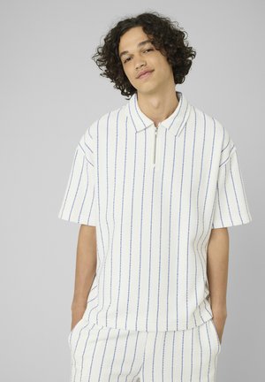 Jeune personne aux cheveux bouclés portant un polo blanc à manches courtes et un pantalon assorti avec de fines rayures verticales bleues, mains dans les poches, fond neutre.