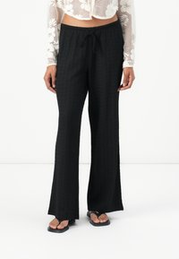 Zwarte, gestructureerde wijde broek met een trekkoord in de taille, met een ontspannen pasvorm. Gecombineerd met zwarte teenslippers.