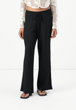 Pantalons larges en noir, texturés, avec une taille à cordon, offrant une coupe décontractée. Associés à des tongs noires.