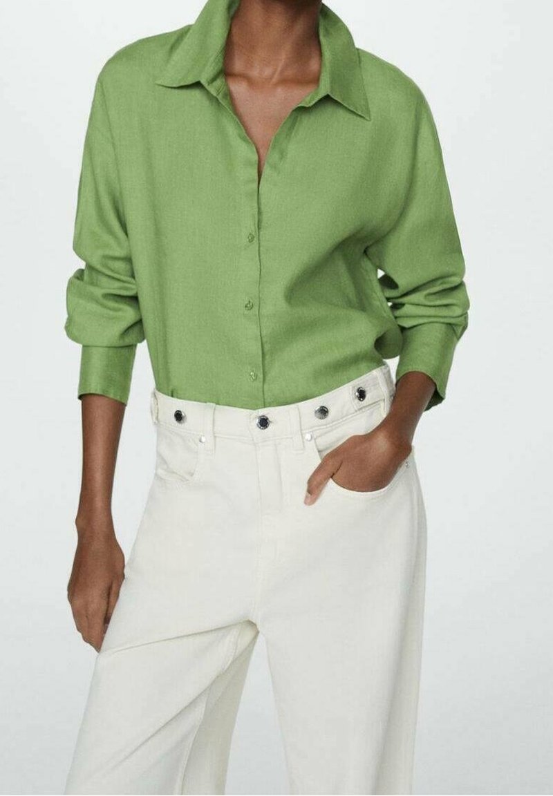 Personne portant une chemise ample vert clair à boutons avec manches retroussées et un pantalon taille haute écru, une main dans la poche.