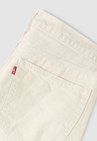 Μπεζ βαμβακερό παντελόνι με πίσω τσέπη που διαθέτει διπλή ραφή και μικρή κόκκινη ετικέτα με λογότυπο κοντά στη μέση.