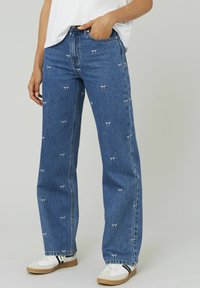 Jean en denim bleu avec des motifs de nœuds blancs brodés. Conception à jambes larges, style classique à cinq poches, fini avec des coutures visibles et un ourlet effiloché.