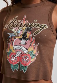 Brun, ärmlös crop top med et fargerikt grafisk motiv av en fugl, dolk, flammer og roser med teksten "Burning wild heart." Laget av bomull.