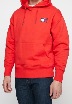 Mężczyzna w jaskrawoczerwonej bluzie z kapturem i kieszenią z przodu oraz logo "Tommy Jeans" na piersi, w ciemnoniebieskich dżinsach.