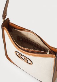Tans en crèmekleurige schoudertas van canvas met leren afwerking. Voorzien van een opvallend logo, ritsluiting en meerdere binnenvakken.