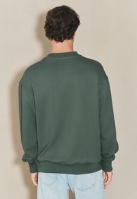 Mørkegrøn sweatshirt med rund hals, ribstrikkede manchetter og kant. Har en afslappet pasform og glat, tekstureret stof. Set bagfra.