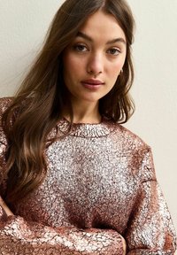 Top de manga larga en oro rosa con un patrón texturizado y agrietado. Cuello de barco y ajuste relajado, con material brillante.