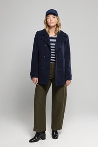 Abrigo de marinero cruzado azul marino, camiseta de rayas, pantalones de pierna ancha verde oliva, botas negras de tobillo y una gorra azul marino, de pie contra un fondo gris.