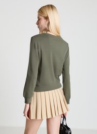 Sweatshirt vert olive à col rond, bas côtelé et manches longues, associé à une jupe plissée beige. Design et texture simples.