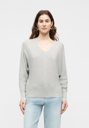 Woman wearing light grey V-neck sweater with subtle ribbed texture and light blue jeans, standing against plain light background.
