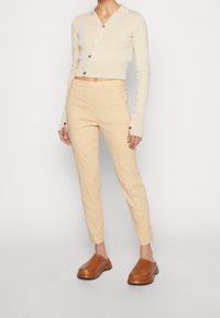 Personne portant un pantalon slim beige, un pull cropped crème à boutons avec des trous pour les pouces, et des sabots marron, se tenant sur un sol gris.