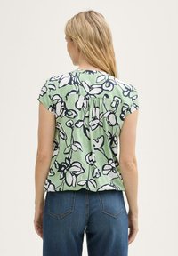 Blouse à imprimé floral avec des manches courtes, base verte, fleurs bleu marine et blanches, coupe décontractée, détail plissé au dos, associé à un jean bleu.