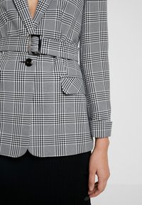 Rutig kavaj i svart och vitt, med bältad midja, enkel knappstängning och en sidoficka med en texturerad tygfinish.