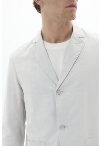 Helle graue Leinenjacke mit drei Knöpfen, Reverskragen und aufgesetzten Taschen. Über einem weißen Rundhals-Shirt getragen.