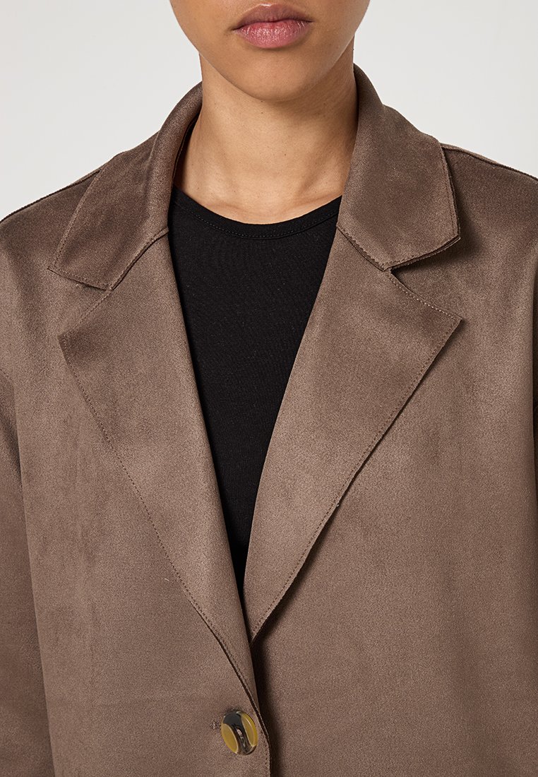 Persona che indossa un blazer in suede marrone sopra una maglietta nera a girocollo, con il colletto, le revers e un solo bottone allacciato.