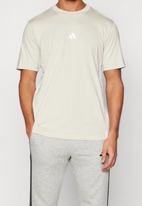 Svetle béžové tričko s krátkým rukávem s bílými bočními panely a malým bílým logem na hrudi. Měkká látka, volný střih.