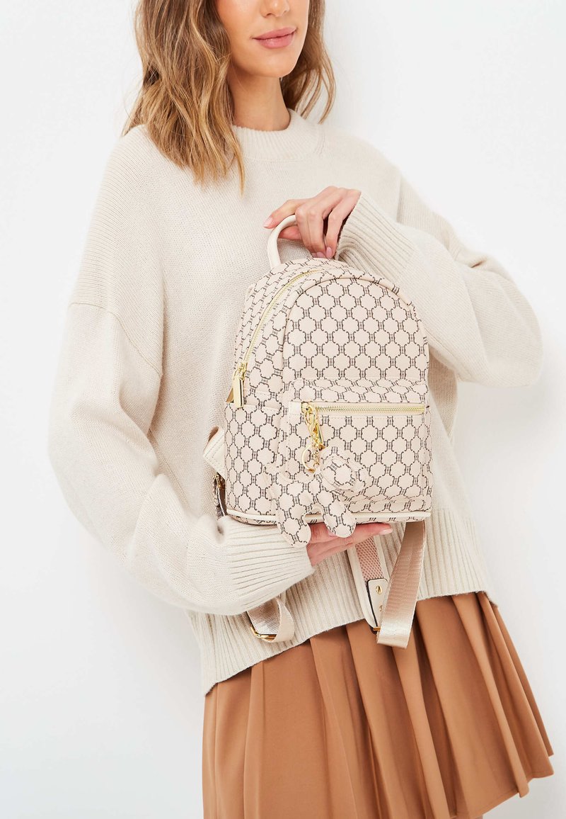 Beige ryggsäck med ett geometriskt mönster, gulddragkedjor och en liten accentcharm. Mjuk material och rundad design med justerbara axelremmar.