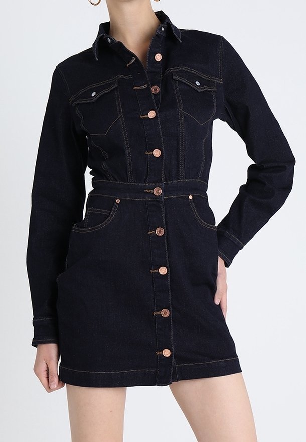 Vestido abotonado de mezclilla oscura con mangas largas, bolsillos en el pecho y detalle de cinturón, llevado por una persona de pie con una mano en la cadera.