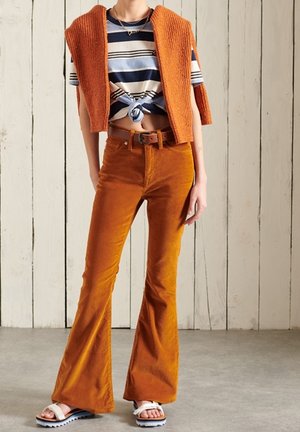 Pantalones de pana acampanados en color naranja quemado, combinados con un top corto a rayas y un chaleco de punto naranja. Sandalias blancas con suelas texturizadas.