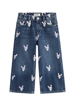 Smilende barn med krøllet hår iført en hvid t-shirt, blå jeans med kaninprint og hvide sneakers, stående mod en ensfarvet baggrund.